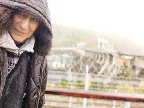 Funny middle-aged adult tourist in jacket during the rain and a hydroelectric power station in the background