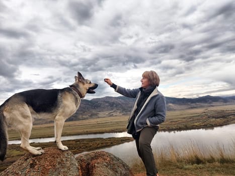 Adult girl with shepherd dog taking selfie near water of lake or river in mountain. Middle aged woman and big pet on nature. Friendship, love and fun