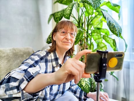 Smiling woman in glasses using a smartphone on a tripod for a video chat. A middle-aged woman posing and taking a selfie