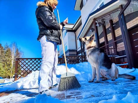 A girl or a woman with a snow broom at home and a large German Shepherd dog. Woman Clearing Snow at Home With Her German Shepherd