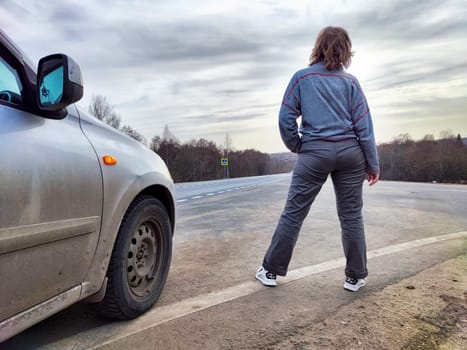 Girl Resting on Road Trip at Dusk by Her Car. A traveler takes a break, leaning on her parked car on an open road, with the sky hinting at dusk