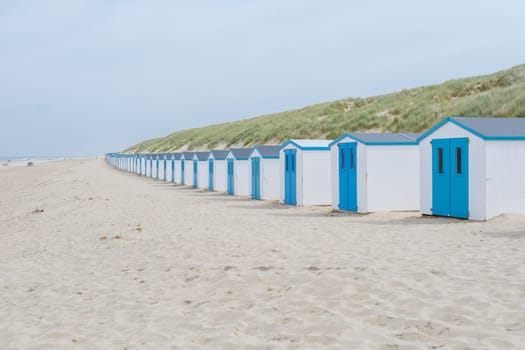A line of charming beach huts with vibrant blue doors standing on the sandy shores of Texel, Netherlands, creating a picturesque and colorful scene. De Koog beach Texel