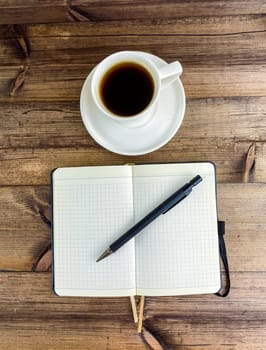 Coffee in a cup and a notepad with a pen, top view. Vertical photo. Coffee in a cup with a saucer and a notebook with a pen on a wooden table.