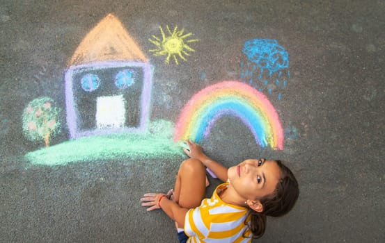 A child draws a house and a rainbow on the asphalt with chalk. Selective focus. Kid.