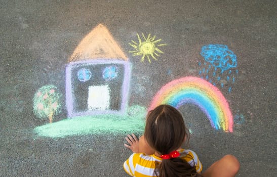 A child draws a house and a rainbow on the asphalt with chalk. Selective focus. Kid.