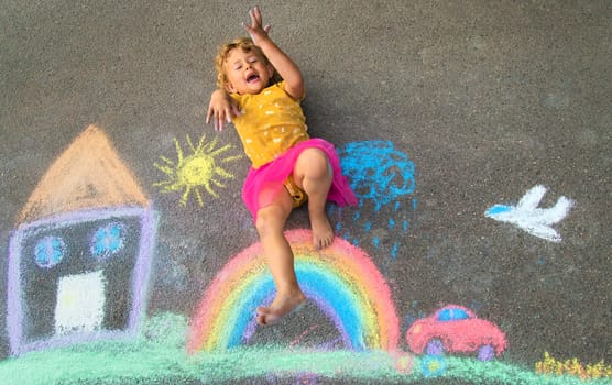 A child draws a house and a rainbow on the asphalt with chalk. Selective focus. Kid.