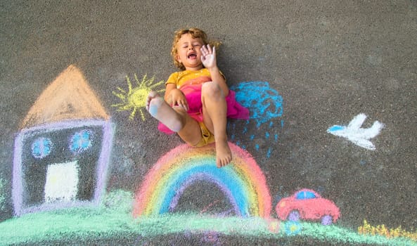 A child draws a house and a rainbow on the asphalt with chalk. Selective focus. Kid.