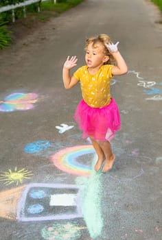 A child draws a house and a rainbow on the asphalt with chalk. Selective focus. Kid.