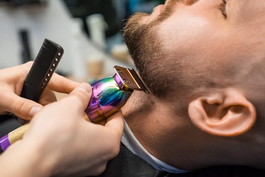 Barber cuts man beard creating stylish shape with trimmer in barbershop closeup. Hairdresser works with male client using machine in salon