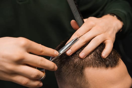 Hairstylist sculpts the brunette hair of the client using scissors at the barbershop