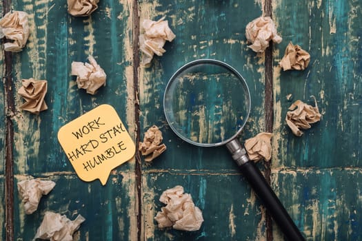 A yellow sticky note with the words work hard, stay humble written on it is placed on top of a pile of crumpled paper. A magnifying glass is also present on the paper, adding a sense of curiosity