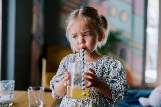 Little girl drinks juice from a glass through a straw while standing at a table in a cafe. High quality photo