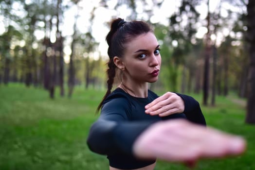 Young woman doing martial arts training in sporty black top holding hands in fighting pose against forest background.