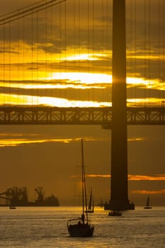 Sailing boat is sailing towards the bridge at bright yellow sunset - oil producing vessel on the background. Vertical shot