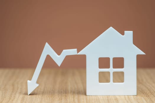 Reducing the interest rate for a mortgage or lowering the cost of insurance for real estate. White house model and graphic arrow pointing down close-up on a brown background. copy space