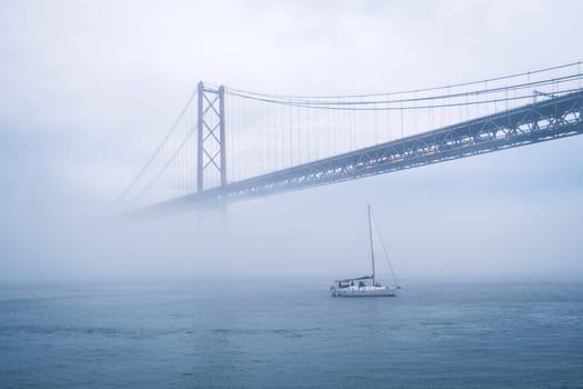 View of 25 de Abril Bridge famous tourist landmark of Lisbon connecting Lisboa and Almada in heavy fog mist wtih yacht boats passing under. Lisbon, Portugal
