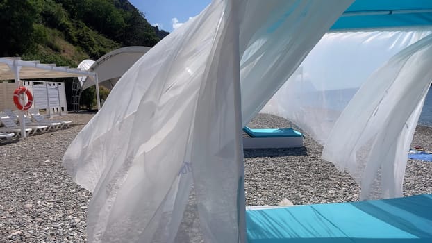 A beach scene with a white tent and blue umbrella. The umbrella is blowing in the wind and the tent is open