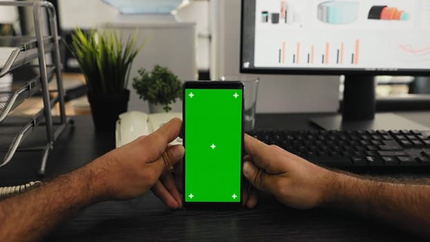 POV of manager holds smartphone with isolated greenscreen layout, working with blank mockup chromakey template at office desk. Young man looking at mobile device display with copyspace.