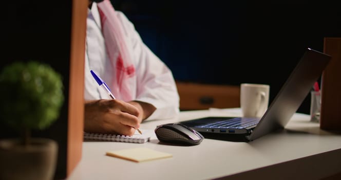 Man in apartment paying attention in elearning seminar, writing on notepad using pen. Middle Eastern homeschooling student listening to lesson while doing his homework tasks, close up