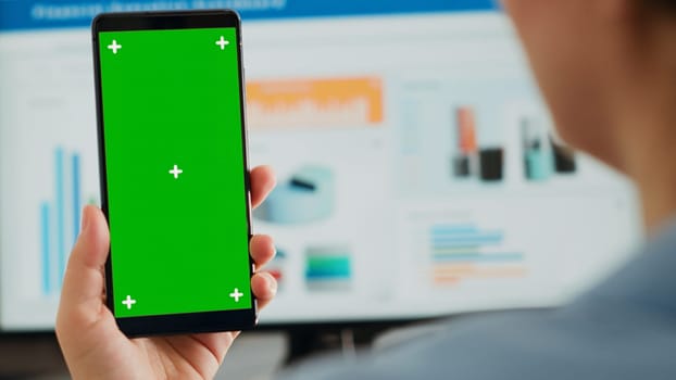 Business person holding greenscreen device at desk, looking at blank mockup display in coworking space. Woman showing mobile phone displaying isolated chromakey template, screen layout. Close up.