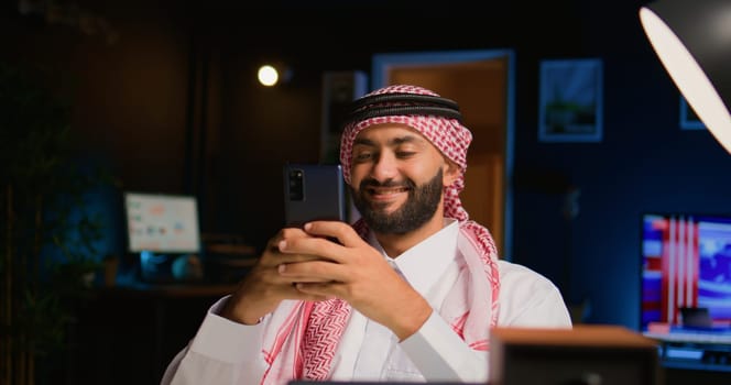 Smiling arab man at home in living room typing messages on smartphone. Middle Eastern person holding cellphone, enjoying relaxing leisure time talking with friends over online messaging app