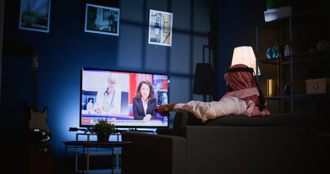 Arabic man lounged on sofa, watching news broadcast while relaxing in living room after hard day at work. Middle Eastern person loafing on couch looking at TV channel in dimly lit apartment