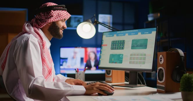 Cheerful muslim broker investor comparing stock exchange valuation financial profit numbers. Arab stockholder in dimly illuminated room looking at market shares growth charts, taking notes