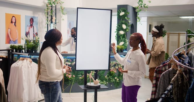 BIPOC shopkeeper in clothing store using augmented reality mockup screen to show woman garment options for her to pick. Worker using led display to help customer visualize her fashion choices