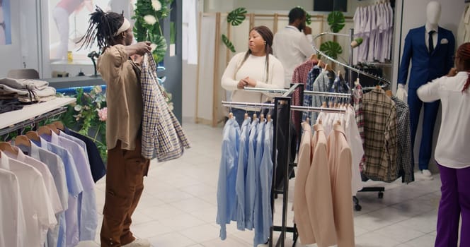 Couple in thrift shop looks at clothes, trying to find cheap but stylish shirts. Woman helping husband choose between garments in second hand clothing store, happy with low price