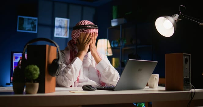 Tired arab businessman in traditional attire suffering migraine working at home office desk. Stressed Middle Eastern worker in living room answering emails getting headache from approaching deadline