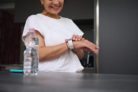 Close-up shot hands of a smiling heathy young woman checking smart wrist watch. A bottle of pure water on the foreground. Fitness app, mobile app. People. Health care and modern wireless technology