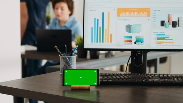 Empty workstation with greenscreen display on mobile phone app, desktop showing marketing analysis and reports in coworking space. Business office desk displaying smartphone screen with chromakey.