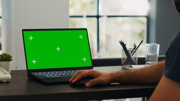 Male employee looks at greenscreen chromakey display while he sits at creative agency workstation, working on e commerce operations. Professional analyst checks blank mockup layout on laptop.