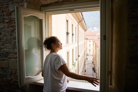 Side view. Smiling happy young woman in white pajamas, opening windows in the living room, contemplating the beautiful touristic city of Como in Lombardy, with red roof tiles of cozy houses. Italy.