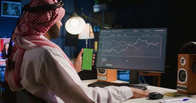 Man holding green screen smartphone while looking at business annual report graphs on computer. Self employed person with chroma key phone in hand performing tasks on device