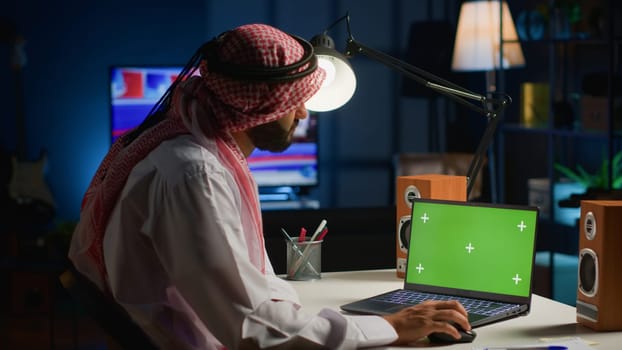 Arab man browsing on mockup laptop while working from home. Self employed freelancer checking emails on isolated screen device in apartment office with TV as background noise