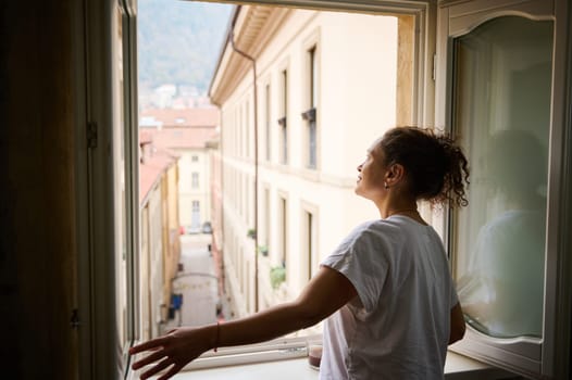 Happy young woman in white t-shirt, opening windows in the living room, enjoying the morning of her holidays, contemplating the beautiful touristic city of Como with red roof tiles of cozy houses