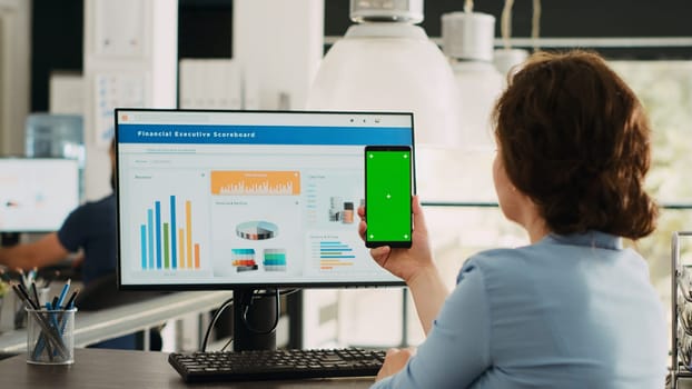 Employee looks at greenscreen on phone, working on business operations at desk. Person holding smartphone device with isolated display showing mockup template, blank layout screen.