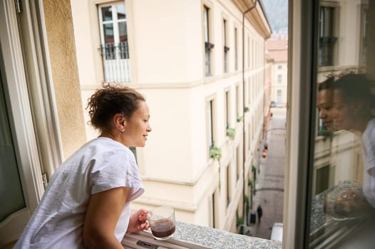 Attractive multi ethnic smiling brunette young woman in white pajamas, enjoying a morning, standing by opened window overlooking the medieval city of Como, expressing happiness and positive emotions