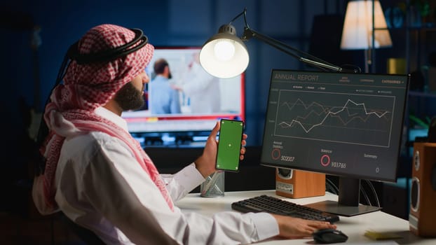 Remote worker holding green screen smartphone while looking at business annual report graphs on computer. Self employed freelancer with mockup phone in hand performing tasks on device