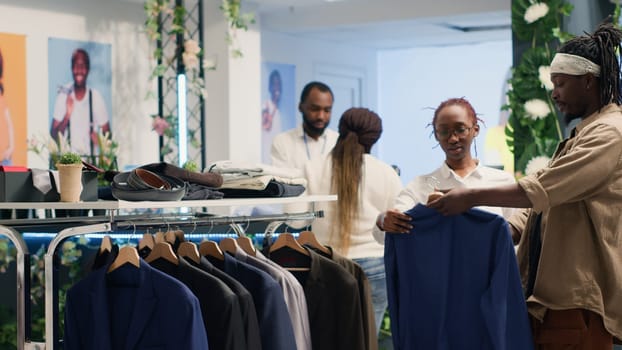 African american man in fancy fashion boutique helped by employee to find suiting stylish formal clothes. Helpful worker assisting client to find fitting designer clothing in premium showroom