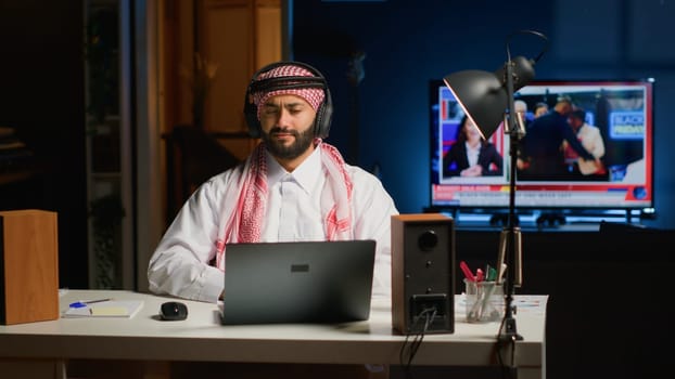 Arab employee with wireless headphones enthusiastic about watching TV show on laptop from living room. Muslim worker doing internet research while listening to music to make time pass faster