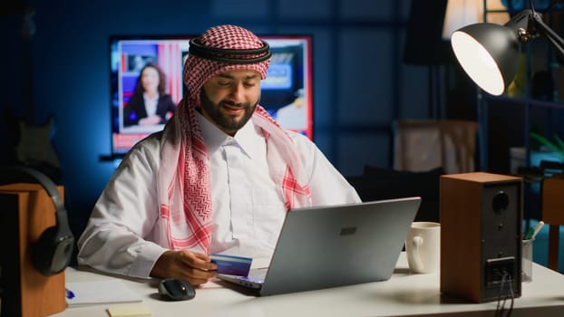 Arabic man doing online shopping, carefully typing credit card information on computer. Muslim person adding payment method on website while in cozy stylish warm home at night