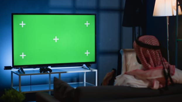 Arab man watching news broadcast on chroma key device while relaxing at home after hard day at work. Muslim person chilling on couch, looking at isolated screen TV channel