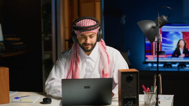 Arab company executive with wireless headphones enthusiastic about working from home office. Smiling Middle Eastern entrepreneur doing online research while listening to music to make time pass faster