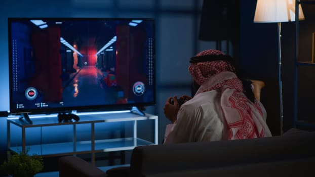 Arab man playing videogames on TV, relaxing after long day at work. Competitive gamer having fun on gaming console in stylish apartment living room, enjoying science fiction shooter game