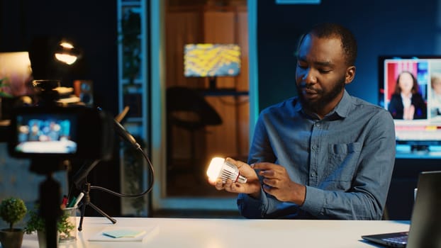 BIPOC content creator in dimly lit living room uses camera to film smart light bulb review for online streaming platforms. Media star hosts internet show, unboxing home automation product