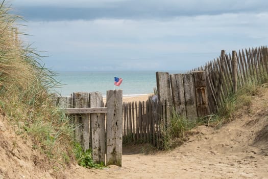 Utah Beach in Normandy, France. At a distance USA flag on wood sea fence, grass and view up sand dunes. Sunny sky light ble clouds. Blue ocean view. High quality photo