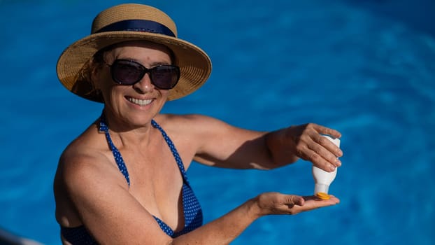 Portrait of an old woman in a straw hat, sunglasses and a swimsuit applying sunscreen to her skin while relaxing by the pool