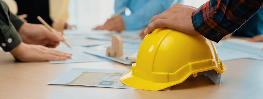 Professional architect team and engineer discuss about architectural project on meeting table with safety helmet, wooden block and architectural document scatter around. Closeup. Delineation.